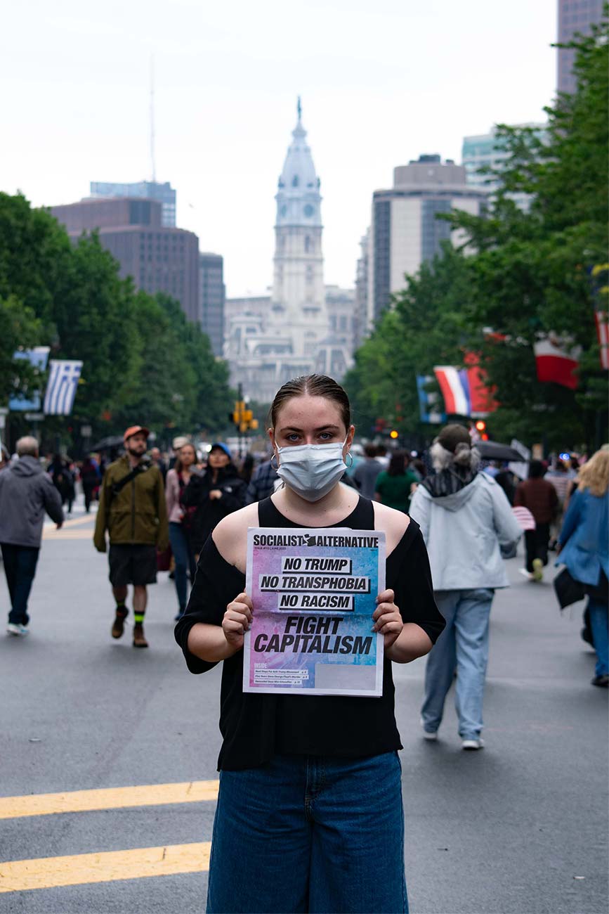 Maya holding a newspaper from Socialist Alternative stating: No Trump, No Transphobia, No Racism, Fight Capitalism. Philadelphia City Hall can be seen in the background